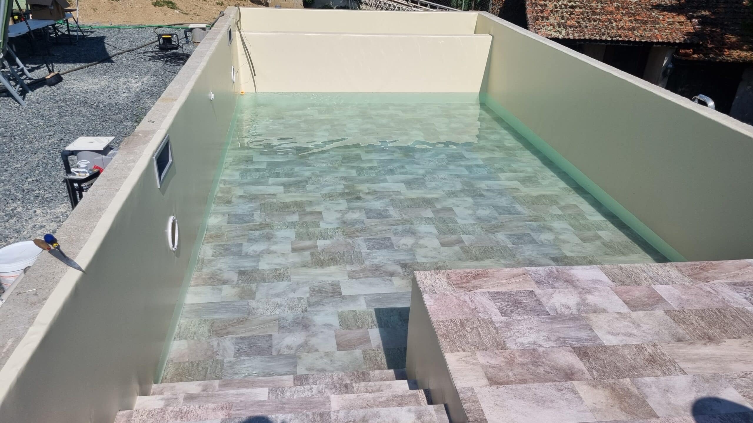 Liner piscine effet carrelage pierre naturelle sur mesure Piscine enterrée en cours de remplissage, montrant un liner beige clair et un fond en carrelage aspect pierre naturelle. Grand escalier d'accès.
