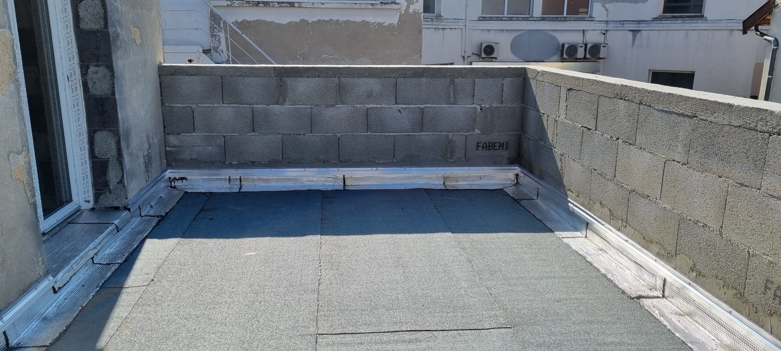 Étanchéité Toit Plat : Membrane, Isolation et Murs FABEMI. Toit-terrasse plat isolé (foil) et étanchéifié par membrane bitumée grise, avec murs en blocs de béton bruts. Profilés de fixation. FABEMI visible.